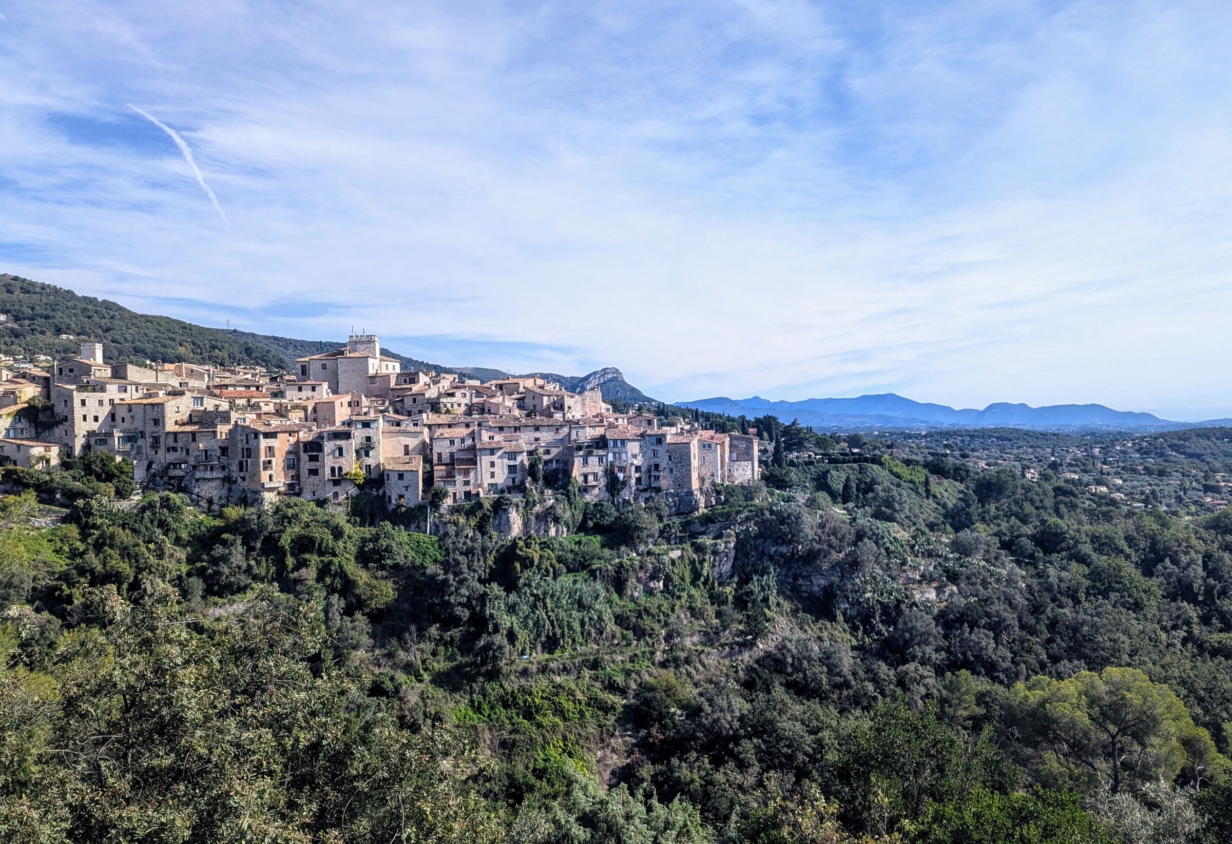 A hilltop village in the south of France at midday, with stone houses on a ridge and mountains beyond